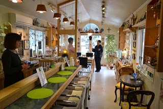 カウンター席や小さなテーブル席が並び、木目調の内装で温かみのある雰囲気のカフェの中で、黒い服を着た客と店員が会話している様子の写真