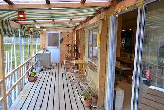 頭上には緑と白の縞模様のオーニングが設置され、木製の床と手すりで囲まれたウッドデッキのテラスに、折りたたみ式の椅子とテーブル、植木鉢などが並び、右側にはガラス戸越しに店内が見えるカフェのテラス席の写真