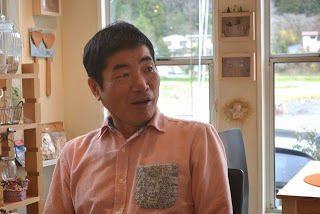 ピンク色のシャツを着た店主の男性が、店内の席に座ってにこやかに話している様子をアップで撮影した写真