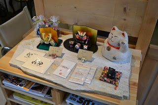 棚の上に招き猫の置物や五月人形風のミニチュア飾り、メッセージカードなどが丁寧に並べられた、和風雑貨のディスプレイの写真