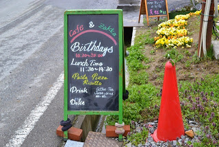 赤い三角コーンと花壇がある店の前の道路脇に置かれた看板に、ランチタイムや提供メニューが記され、店名の「Cafe & Zakka Birthdays」が色とりどりに手書きされている写真
