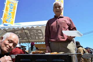 屋外の焼き台の前で、左側に座ってホタテを食している男性と、もう一人の男性が白いトレーを持ってホタテを焼いている様子を写した写真