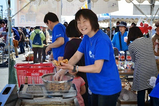 テントの下のフライヤーの前に立つ笑顔の青いTシャツを着た紺野禎子さんが、メッシュボールに入った薄いオレンジ色の海産物の様な物を手に持っている様子を写した写真