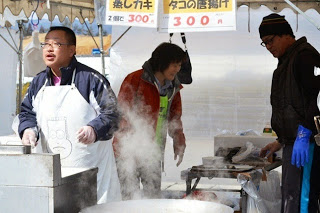 「タコの唐揚げ」などと書かれているテントの下の蒸気の上がる大きな鍋の前でエプロン姿の男性たちが調理をしており、中央に赤色の上着を羽織った女性が立っている写真