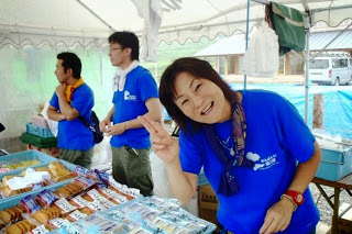 テントの中で2人の男性スタッフが並んで商品の販売をし、隣に立つ青いTシャツの紺野禎子さんが笑顔でピースをしている写真