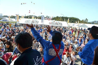 白いテントが立ち並ぶ屋外のイベント会場で、大勢集まった人々に向かって、背中に「福興市」の文字が入った青いジャンパーを着た3人のうちの1人が餅まきをしている後ろ姿の写真