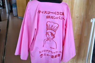 背中にキャラクターと「パティスリーくりこ& 絆カンパニー」と書かれたピンク色の法被がハンガーにかけられている写真