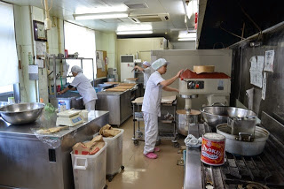 作業台に大きな調理器具や材料が並んでいる厨房の中で、2人のスタッフが作業をしている製造風景の写真