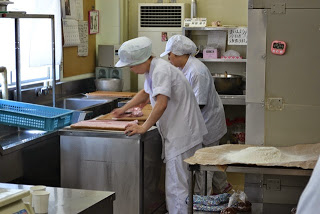 厨房内で白衣と帽子を着た2人のスタッフが、ロールケーキの生地を広げて作業している様子を横から撮影した写真