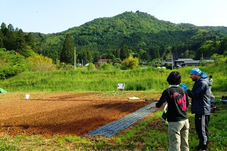 新しく耕された畑の前で、2人の人物が山を背景に話をしており、手前には黒いビニールシートや道具が置かれている畑作業の準備風景の写真