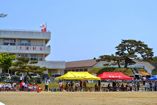 校庭の向こうに「大運動会」と書かれた白い校舎と赤・黄・青のテントが並び、観客や児童が集まっている運動会の様子の写真