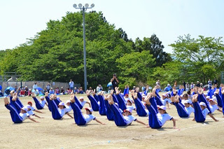 白い帽子と白と青い体操服の児童たちが、地面を両手で支えてお尻をつき、両足を揃えて真っすぐに上げV字バランスを行っている写真