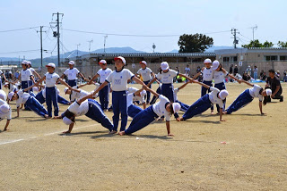屋外のグラウンドで、青いジャージと白い帽子をかぶった児童たちが3人ずつ横一列に並び、真っすぐ中央に立つ児童の両手を、両側の児童が片方の手で握り、もう片方の手を地面について姿勢を保つ組体操の扇を披露している一場面の写真