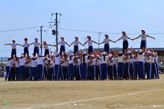 グラウンドの上で複数の児童が下で支え、上段の児童たちは手をつないでバランスを取りながら立っている大規模な組体操のフィナーレのような場面の写真