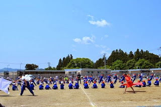 青い体操着の児童たちがグラウンド中央に並び、白と赤の旗を持ったリーダーの合図に合わせて綱引きを行っている写真