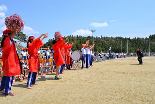 赤い法被を着た応援団の児童が、ポンポンや太鼓を使って元気に応援している写真