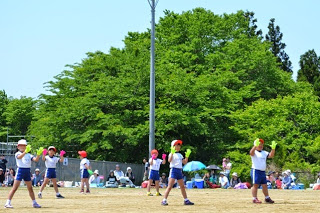 白い体操着を着て両手に黄緑やピンクの手袋をはめた児童たちが、グラウンドで間隔をあけて立ち妖怪体操を踊っている写真