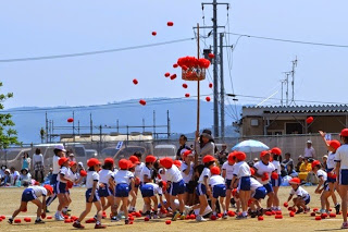 周囲で保護者が見守る中、赤い帽子をかぶった児童たちが、赤い玉を上のカゴに投げ入れている玉入れの様子の写真