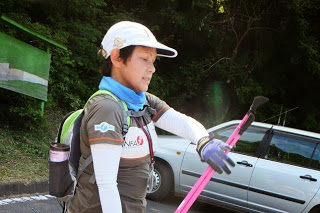 白い帽子をかぶり、首に青いタオルを巻いた女性が、ピンク色の登山用ポールを持っている様子をアップで撮影した写真