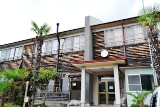 木造二階建ての建物で、外壁には丸い時計が取り付けられ、入口には赤い庇とコンクリートの柱があり、周囲にはヤシの木のような南国風の植物が植えられている旧校舎の外観の写真