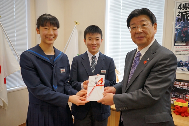 スーツ姿の佐藤町長が右側に立ち、中央の男子学生と左側の女子学生が並んで三人で笑顔を見せながら、紅白の水引が付いた目録を両手で持っている写真