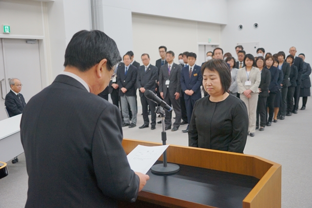 スーツ姿の大勢の人々が整列する中、壇上で町長がマイクの前に立つ女性に向けて辞令を読み上げ、退職者辞令交付式が行われている写真