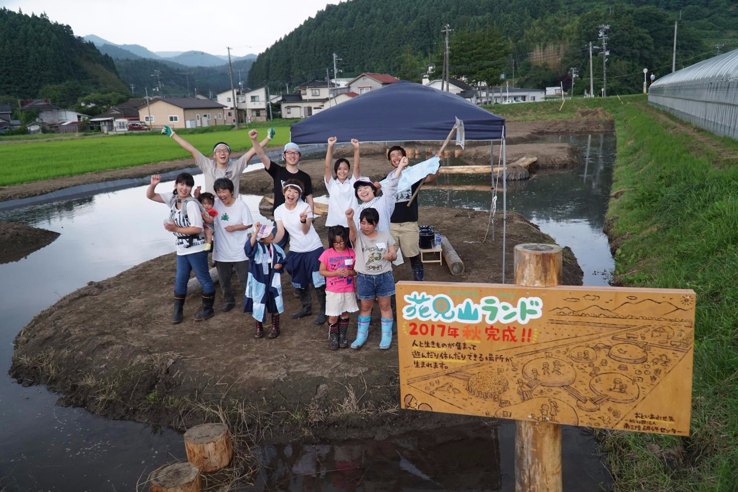 造成されたばかりの花見山ランドの前で、完成を祝って手を挙げて喜ぶ地域住民や子どもたちの集合写真で、前景には「2017年秋完成」と書かれた手作りの看板が立っている写真
