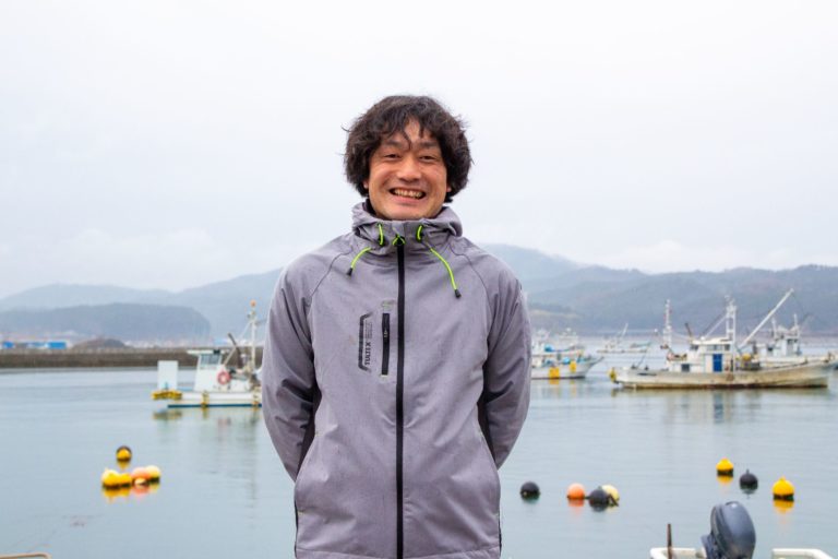 漁船が停泊している港の前で灰色のジャケットを着た後藤新太郎さんが立っており、背景には山々と曇り空が広がっている写真