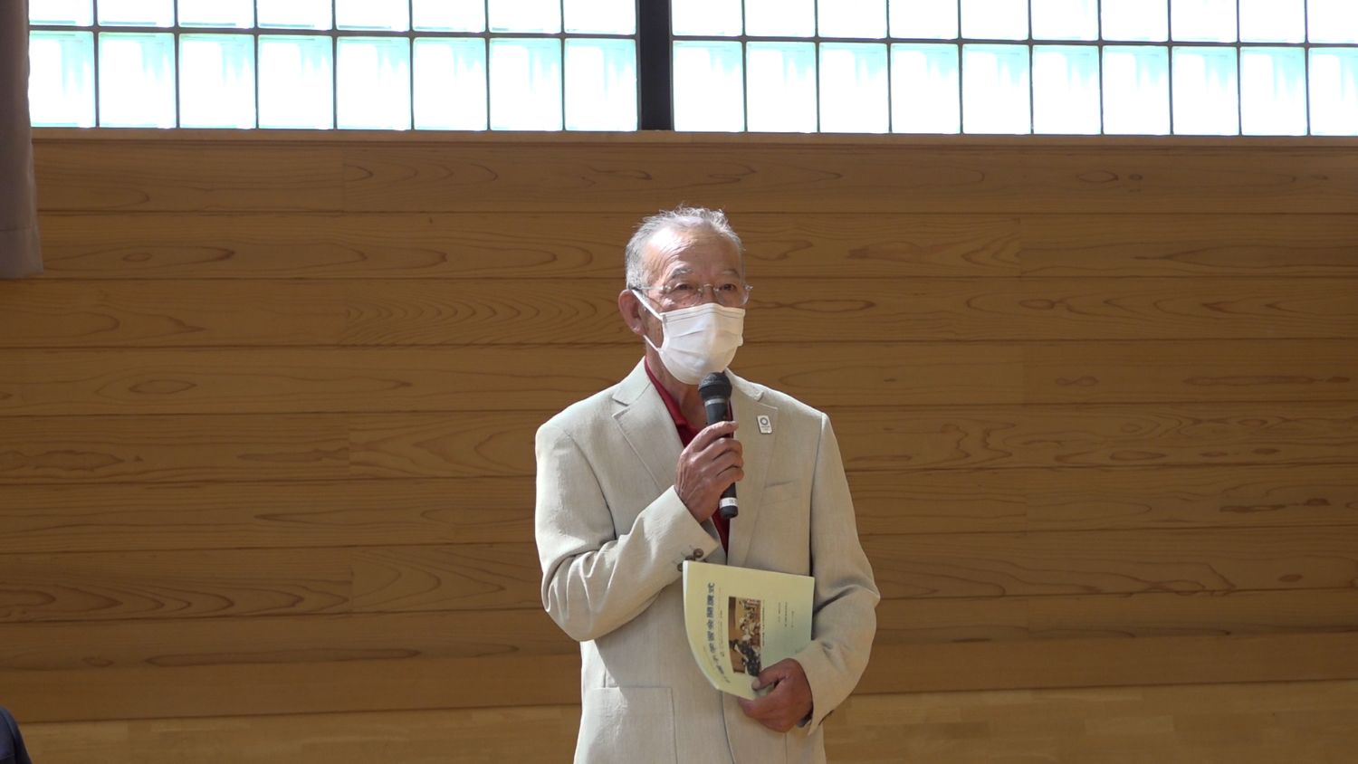 白いジャケット姿の高齢男性がマスクをつけてマイクを持ち、手にパンフレットを抱えて話しており、地域の伝統文化について語っている写真