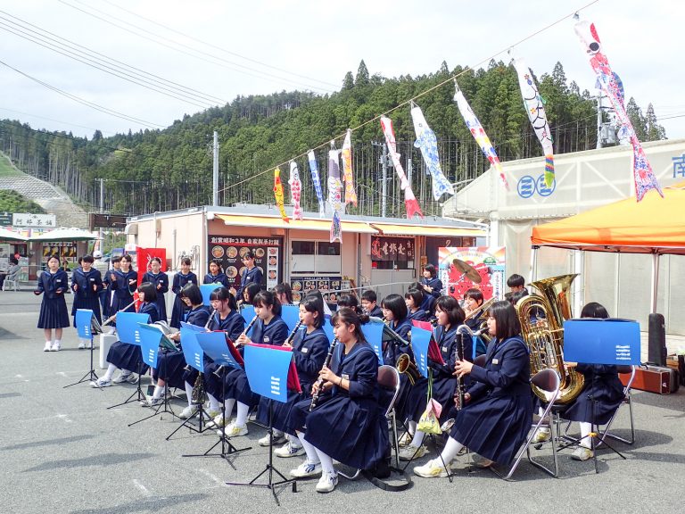 のぼり旗や鯉のぼりが掲げられているさんさん商店の広場で、志津川中吹奏楽部の生徒たちが楽器を演奏している、チャリティーコンサートの写真