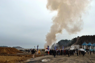 多くの人が集まる屋外の空き地で、焚かれた炎から大きな煙が空高く立ちのぼっている護摩の様子をやや離れた位置からとらえた写真