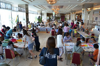 広い室内の会場で、多くの大人や子どもたちがテーブルに分かれて座り、工作や体験学習を楽しんでいる様子の写真