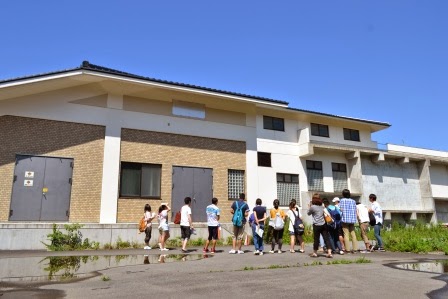 青空の下で十数人の若者たちが、白と茶色の外壁の建物の前に集まり見学している写真