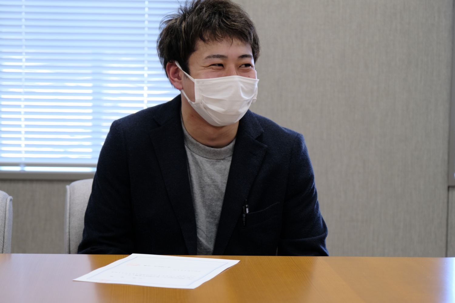 紺色のジャケットと白いマスクをつけた佐藤さんが、室内の木製のテーブルの前に座って笑顔で話をしており、机の上には白い書類が置かれている写真