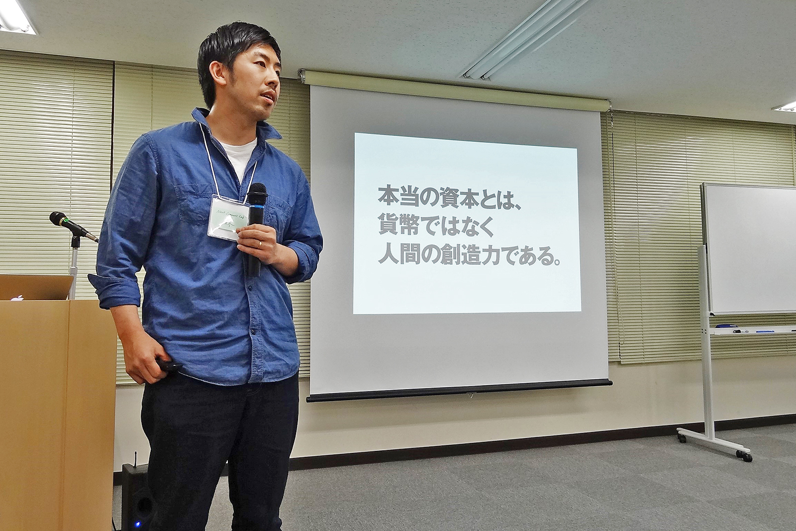 「本当の資本とは、貨幣ではなく人間の創造力である。」と書かれたスライドが映ったスクリーンの横で、マイクを持って立っている林さんの写真