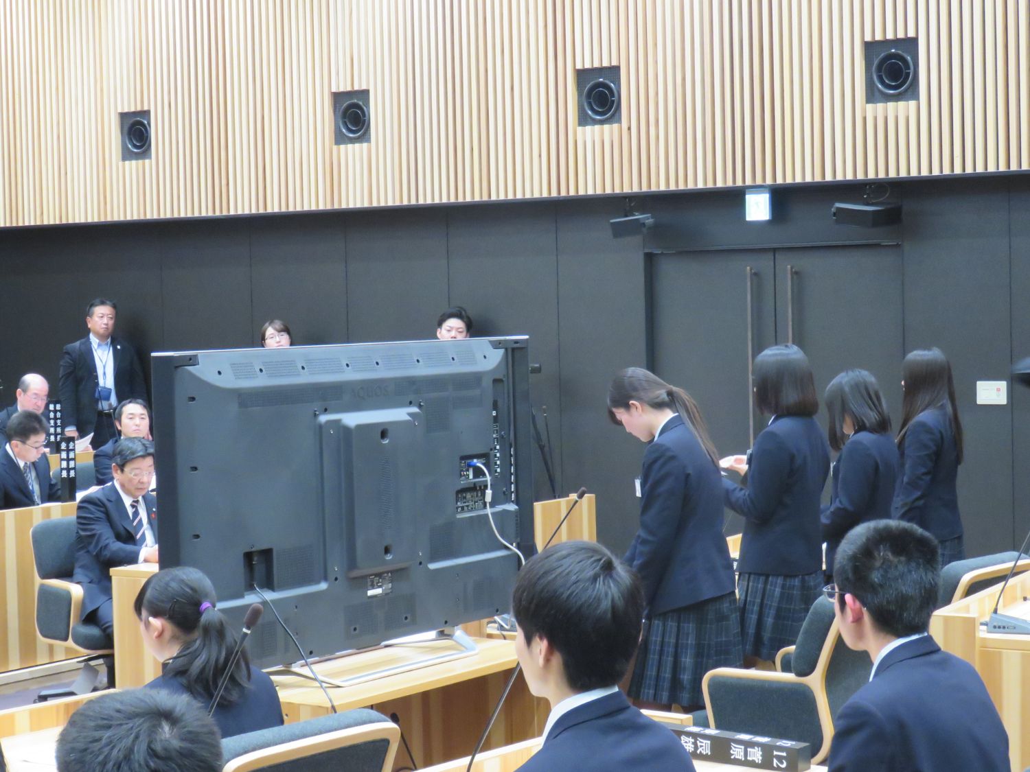 前方の議員席にいる4人の学生が立ち、他の学生や大人たちが椅子に座ってその様子を見守っている様子の写真