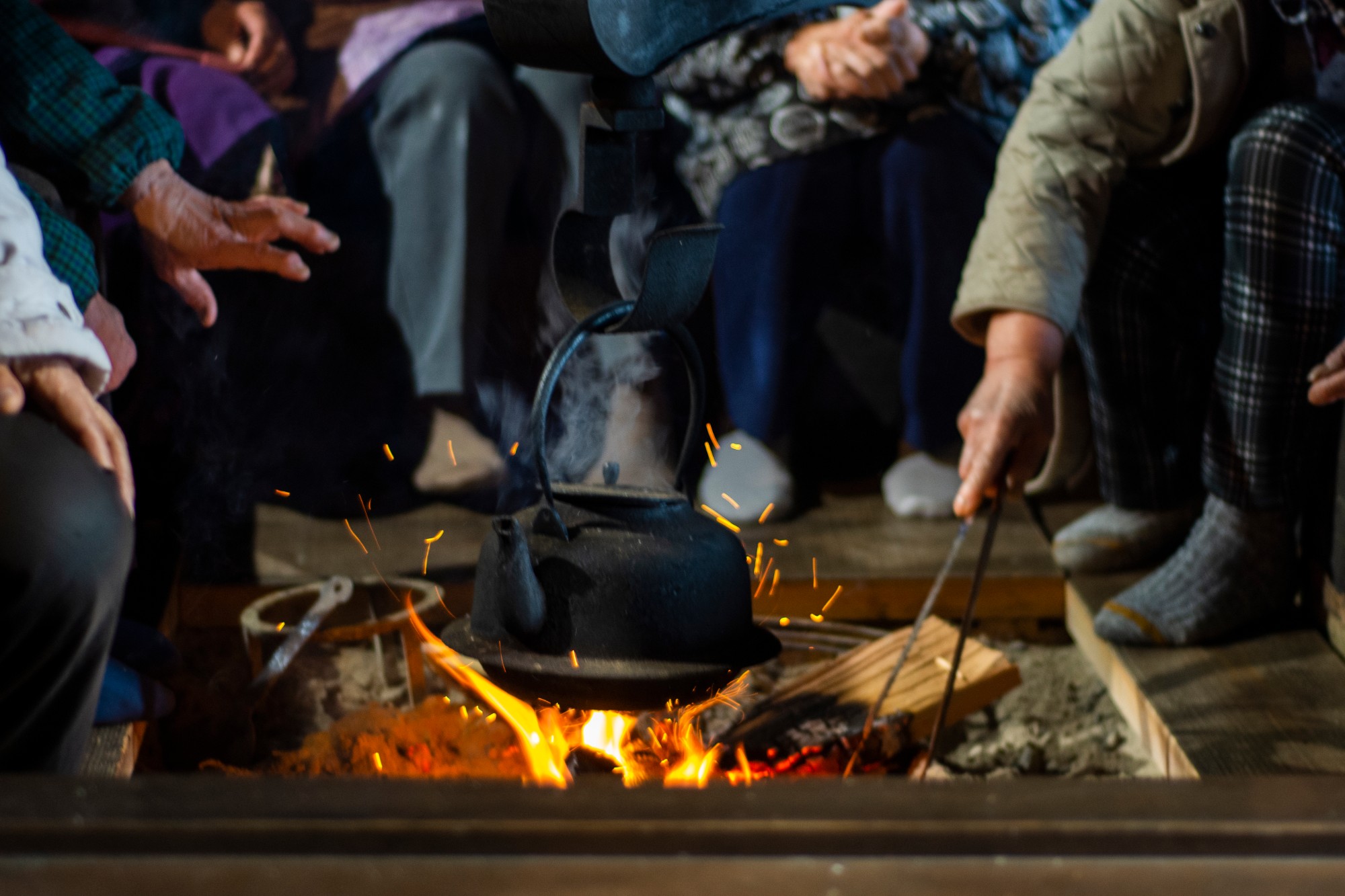囲炉裏を囲んで数人が座り、中央の火に鉄瓶をかけて湯を沸かしている様子が写っている写真