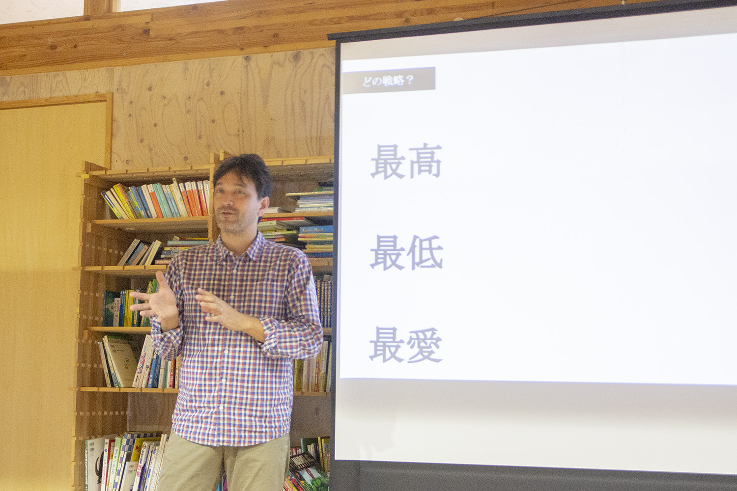チェック柄のシャツを着た男性が、木製の本棚の前で日本語のスライドを使って話している写真