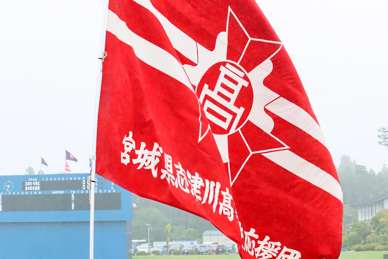 スコアボードと複数の旗が立っているスタジアムが見える背景に、赤地に2本の白の横線が入り「宮城県志津川高等学校応援団」と白文字で書かれた旗が風にはためいている、志津川高校応援旗をアップで撮影した写真