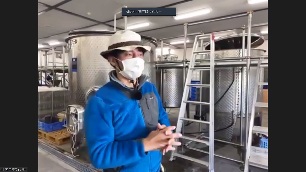 ワインの醸造所と思われる場所で、マスクと帽子を着けた青い服の男性がステンレス製のタンクの前で話しをしているZoom画面の写真
