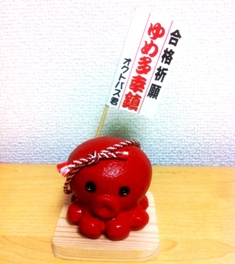 赤いタコの置物が木製の台座に乗っていて、頭には白と赤の紐が巻かれ、背中から「合格祈願 ゆめ多幸鎮 オクトパス君」と書かれた旗が立っているオクトパス君の置物の写真