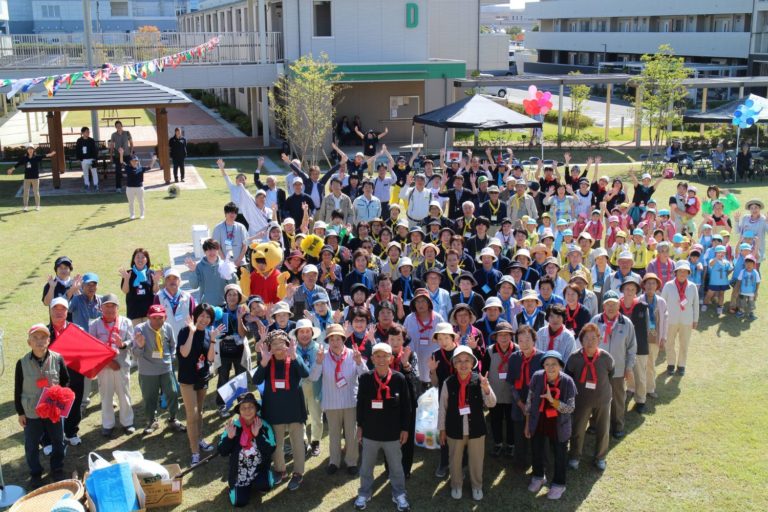 晴天の下、走らないミニ運動会に参加する子供と大人たちが芝生の上に集まり、上空から撮影された集合写真