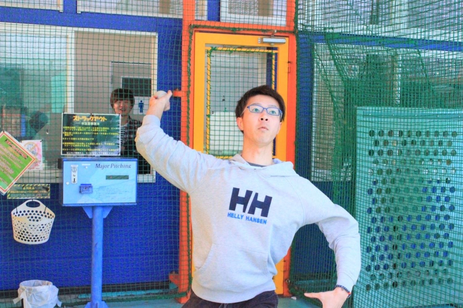 スポーツ施設の一部と思われる場所で、グレーのパーカーを着た男性が腕を上げて何かを投げようとしているようなポーズを取っている写真