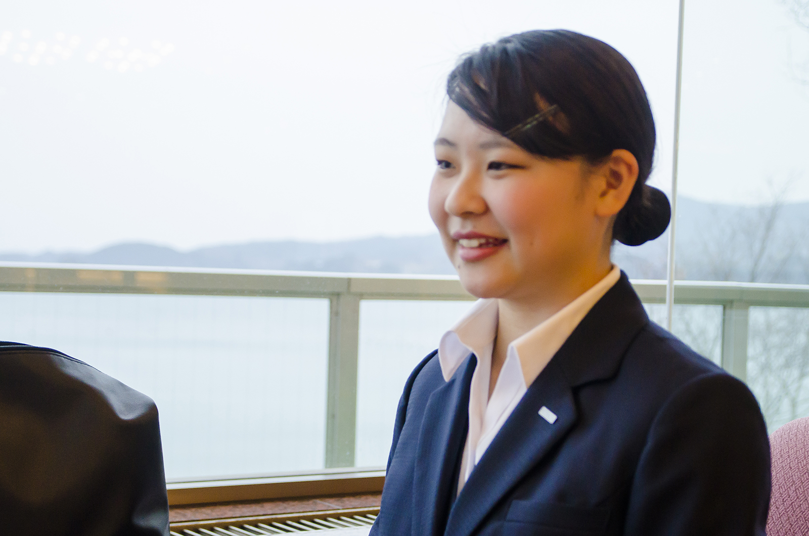 窓辺でスーツ姿の沖澤鈴夏さんが横を向き、柔らかく微笑んでいる写真
