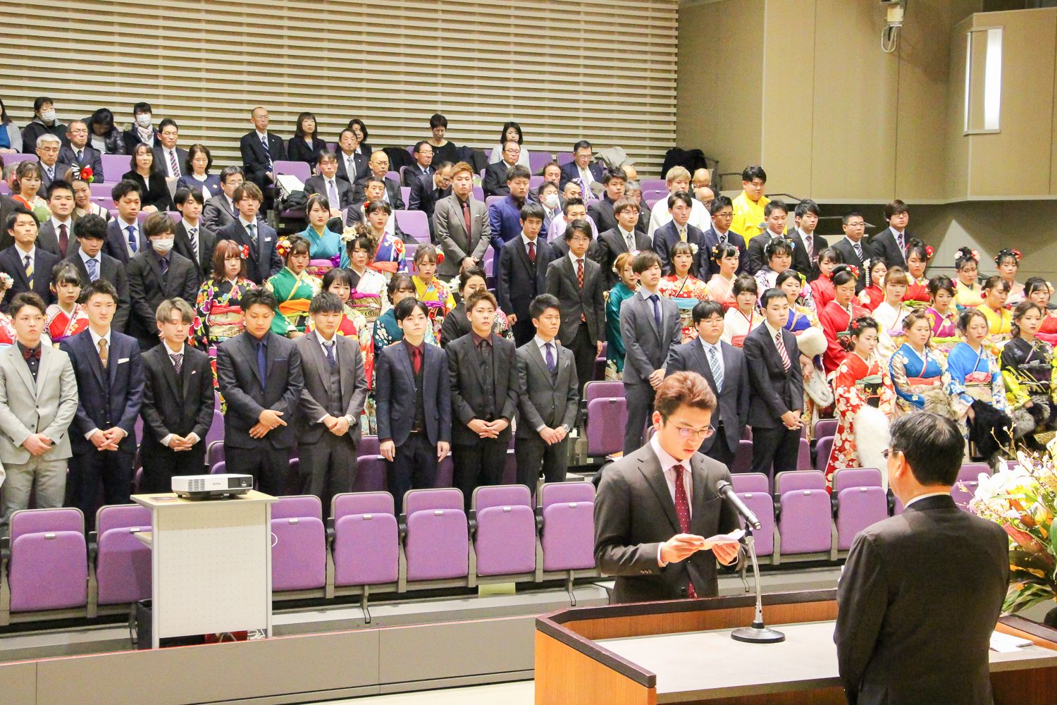 色とりどりの振袖やスーツ姿の成人者達が後ろに整列し、代表者の阿部怜哉さんが誓いの言葉を述べている写真