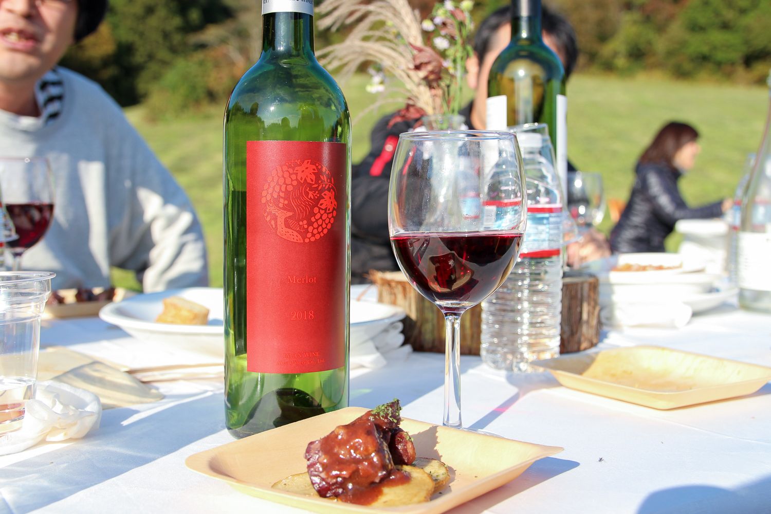 中央には赤いラベルのワインボトルがあり、その横にグラスに注がれた赤ワイン、手前の皿には、赤ワインソースがかかった肉料理が盛られている写真