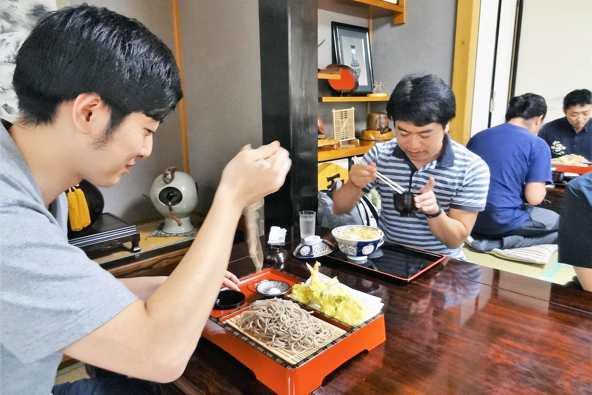 和風の座敷で男性たちが畳に座って昼食をとっており、手前にはざるそばと天ぷら、奥には丼ものを食べる姿がある食事風景の写真
