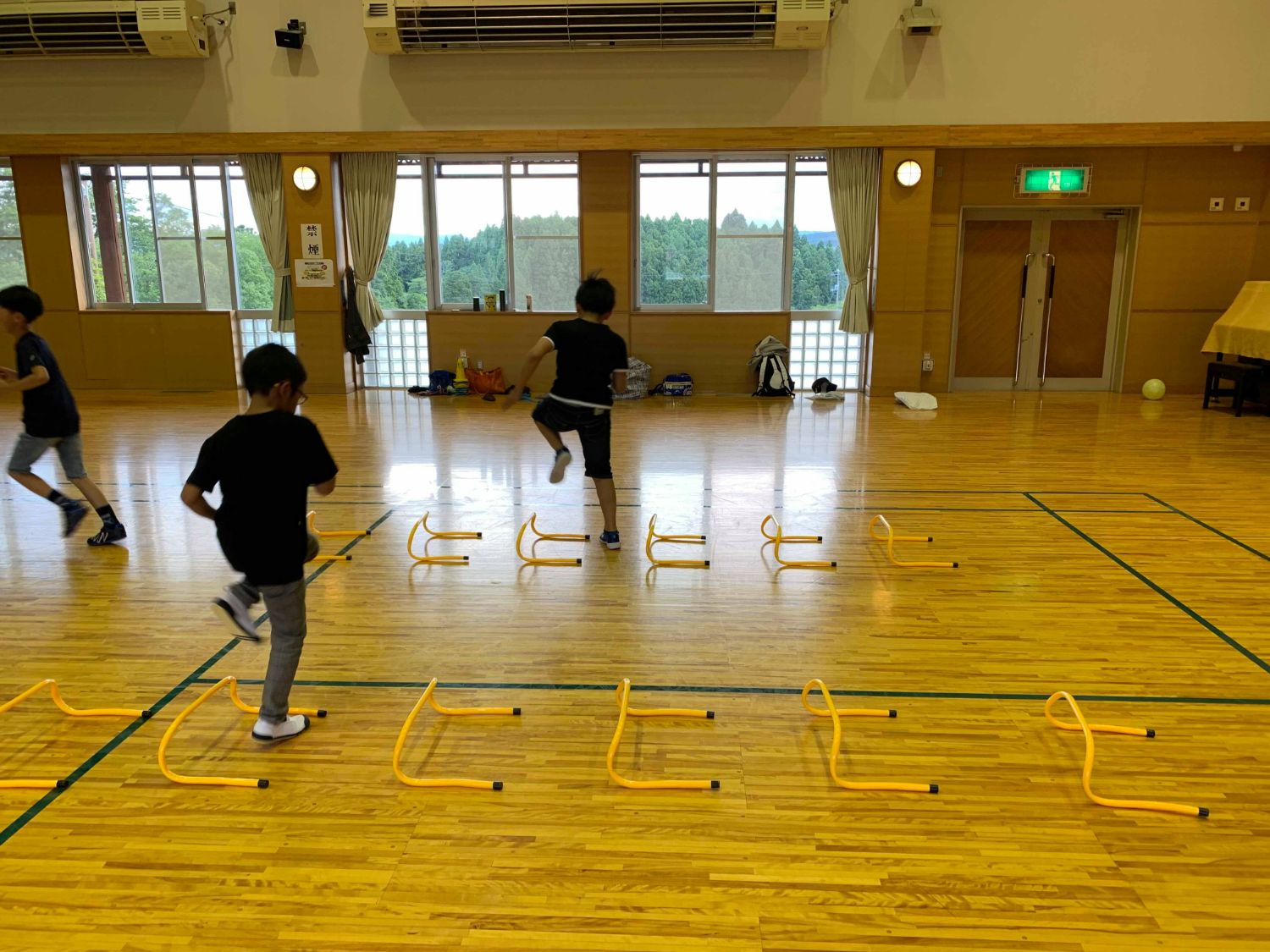 体育館の中に2列に並んだ黄色い小さなハードルを跳び越えて運動する子どもたちの写真