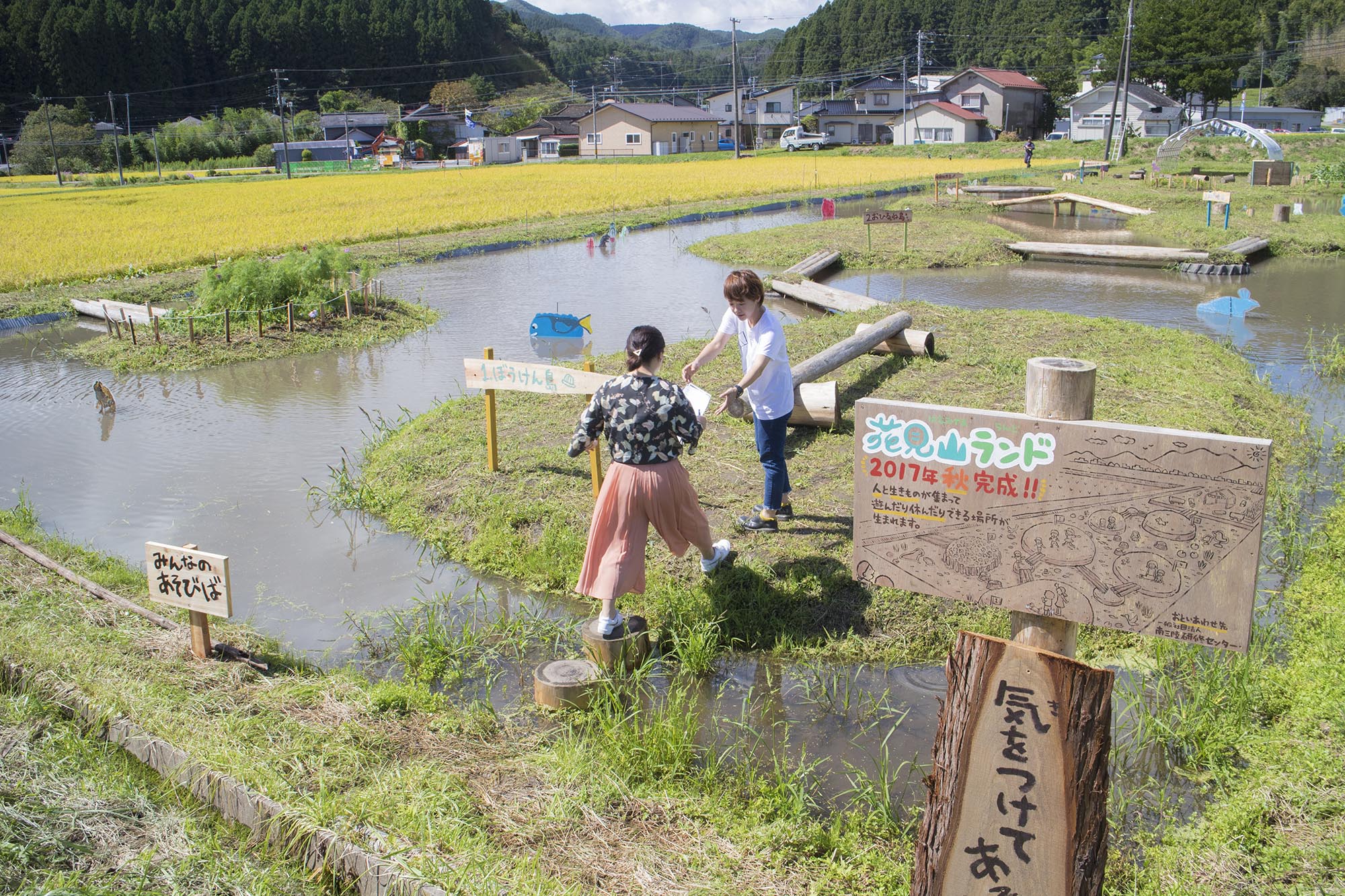「花見山ランド」で、飛び石を渡って水辺の遊び場を進む2人の女性の後ろ姿と、手前に設置された園内案内板が写っている写真