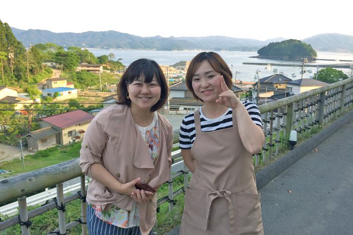 遠くに山並みが見える海辺の住宅地を見下ろす高台で、女性リポーターとベージュのエプロンをつけた女性が並んで笑顔で立っている写真